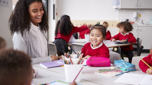 Female teacher and Chinese schoolgirl sitting at a table in an infant school class smiling to other kids, selective focus