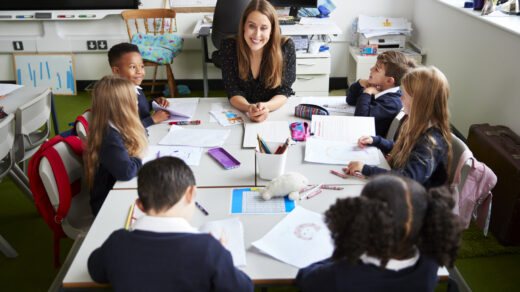 Elevated view of female primary school teacher sitting at a table smiling in a classroom with schoolchildren during a lesson
